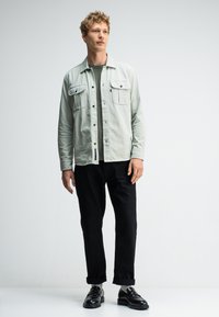 Lichtgroene button-up blouse met twee borstzakken, gecombineerd met zwarte omgeslagen jeans en zwarte loafers, staande tegen een effen achtergrond.