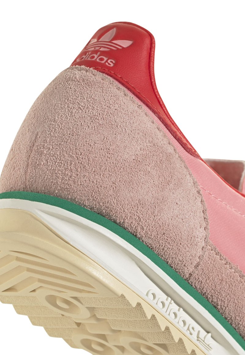 Les baskets Adidas présentent un dessus en suède rose avec un talon en cuir rouge, des accents verts et une semelle texturée blanc cassé.