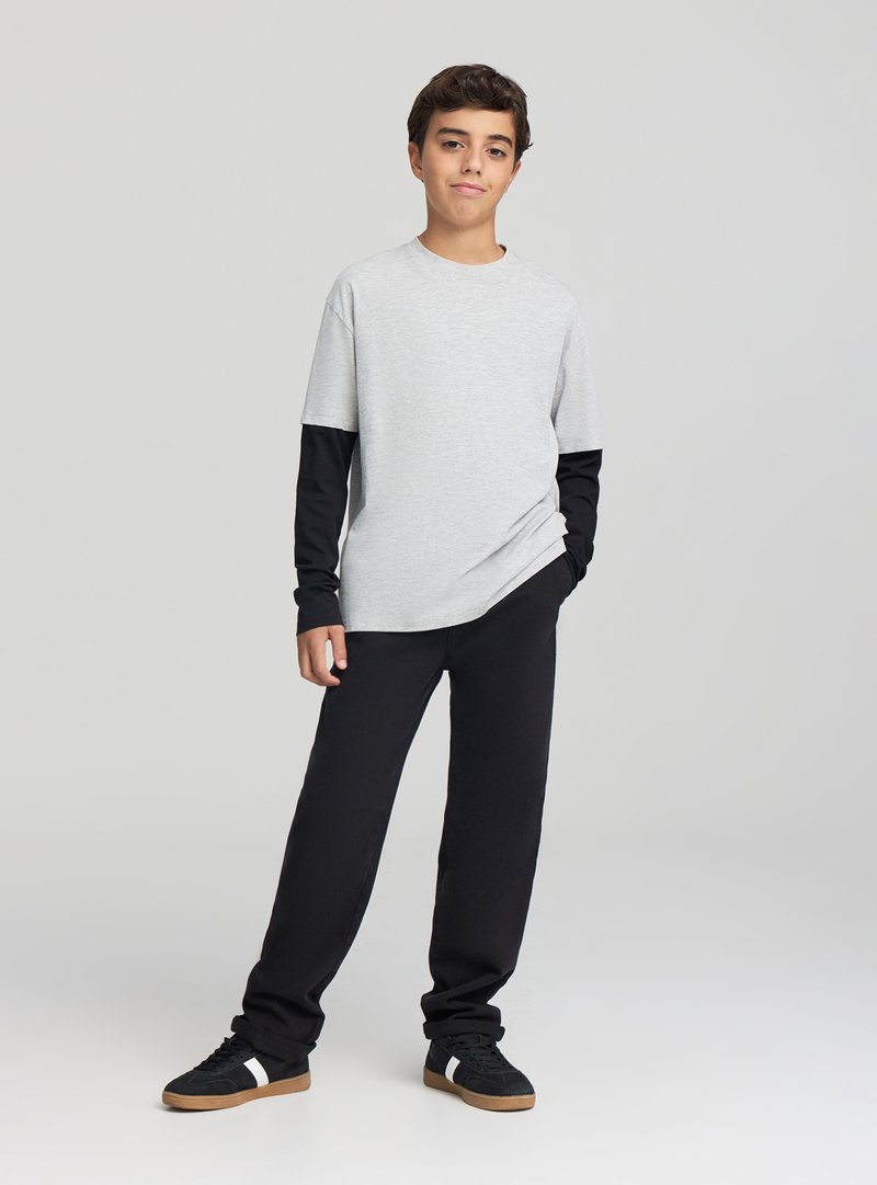 T-shirt grigia a maniche corte indossata sopra una maglia nera a maniche lunghe, abbinata a pantaloni della tuta neri e scarpe nere con dettagli bianchi.