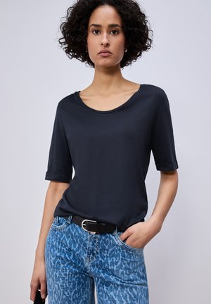 Frau mit lockigem Haar, die ein schwarzes kurzärmeliges Oberteil, blaue Jeans mit Leopardenmuster und einen schwarzen Gürtel trägt, stehend mit einer Hand in der Tasche.