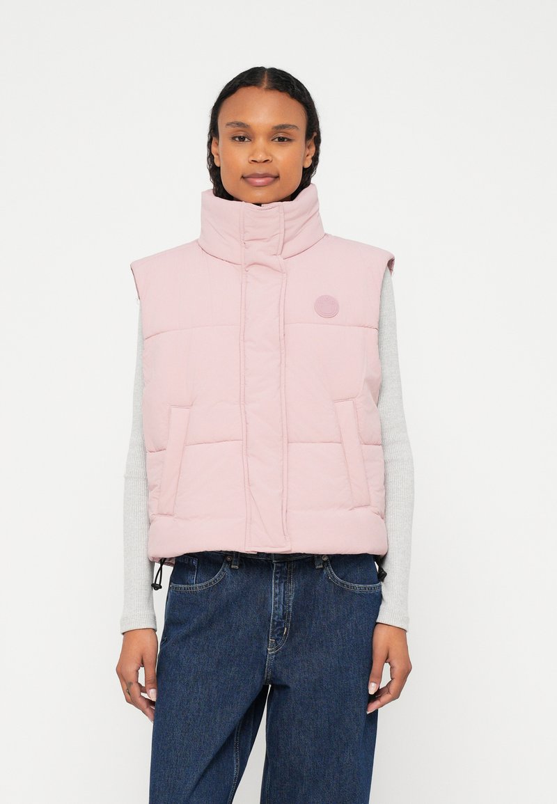 Světle růžová prošívaná vesta s vysokým límcem, dvěma předními kapsami a kulatým logem. Nošena přes světle šedý top s dlouhým rukávem.