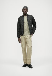 Schwarze gesteppte Bomberjacke, hellgrünes T-Shirt, beigefarbene Cargo-Hose mit Taschen und schwarze Schuhe. Modell trägt eine graue Kappe.