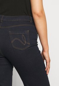 Gros plan d'une personne portant un jean bleu foncé avec des coutures décoratives jaunes sur la poche arrière et un haut noir côtelé.