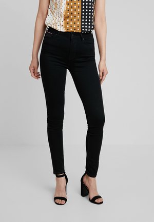 Vrouw draagt zwarte skinny jeans, een mouwloze blouse met patroon en zwarte sandalen met hoge hakken, staande tegen een effen grijze achtergrond.