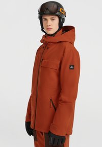 Oranje skijas met een capuchon, voorrits, borstzak en zijrits. Zwarte logo patch op de mouw. Ggedragen met handschoenen en een helm.