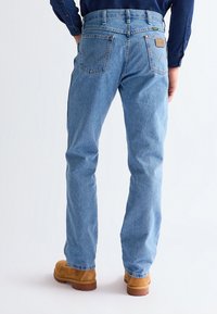 Man wearing light blue straight-leg jeans and tan leather boots, standing with hands in pockets against white background.