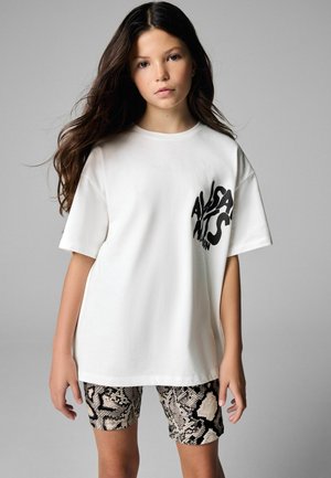 Fată tânără cu păr lung și întunecat, purtând un tricou alb larg cu text negru și pantaloni scurți cu model șarpe, pe un fundal gri.