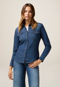 Camicia in denim, blu scuro, maniche lunghe, due tasche sul petto, dettagli in metallo argentato, abbinata a jeans blu chiaro con dettagli borchiati.