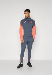 Grå atletisk träningsdräkt med neonorange accenter. Jackan med dragkedja har en hög krage och tre ränder; tajta byxor med matchande randdetaljer.