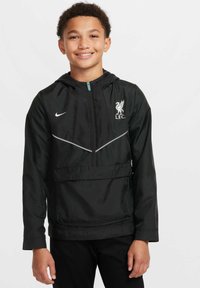 Czarna wiatrówka Nike z kieszenią na przodzie, kapturem i logo Liverpool FC. Posiada odblaskowy szary pasek na klatce piersiowej. Gładki, lekki materiał.