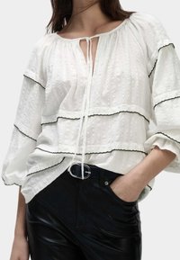 Blusa blanca texturizada con ribete negro, con un diseño de cuello atado y volantes en capas. Usada con pantalones negros, mostrando un cinturón elegante.