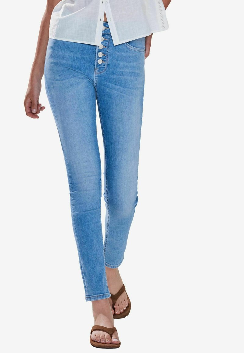 Vaaleansiniset skinny farkut korkealla vyötäröllä, joissa on viiden napin etulukitus ja vähäistä kulutusta. Yhdistetty ruskeisiin sandaaleihin.