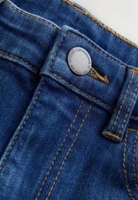 Denim jeans in een donkerblauwe kleur, met gouden stiksels en een metalen knop met "NEXT AUTHENTIC" erin gegraveerd. Close-up detail.