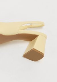 Beige slingback-kengät matalalla, paksulla korolla ja ommellulla joustoresorivillä valkoista taustaa vasten.
