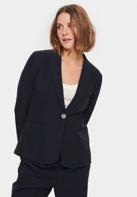 Jūras spēku zila bleizeris ar vienu pogu, ar izgrieztiem atlociem un divām priekšējām kabatām, izgatavots no gludas, teksturētas auduma. Kombinēts ar bēšas krāsas topu.