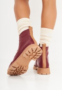 Botas de tobillo de cuero burdeos con detalles en marrón claro, suela de goma texturizada y diseño de cordones. Calcetines de crema acanalados visibles por encima de las botas.