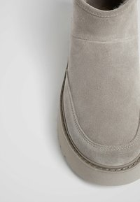 Grå mocka ankelsko med rund tå, slät yta, detaljer med sömmar och tjock sula. Ovandelen har en minimalistisk design och rena linjer.