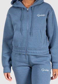 Sudadera azul con cremallera y cordones, con puños y cinturilla de canalé, y un logo blanco bordado en el lado izquierdo del pecho. Tejido suave.