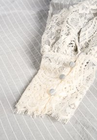 Krajková blúzka so zapínaním na manžetách v off-white farbe s komplexnými vzormi, s tromi šedými gombíkmi, na pozadí šedého pruhovaného materiálu.