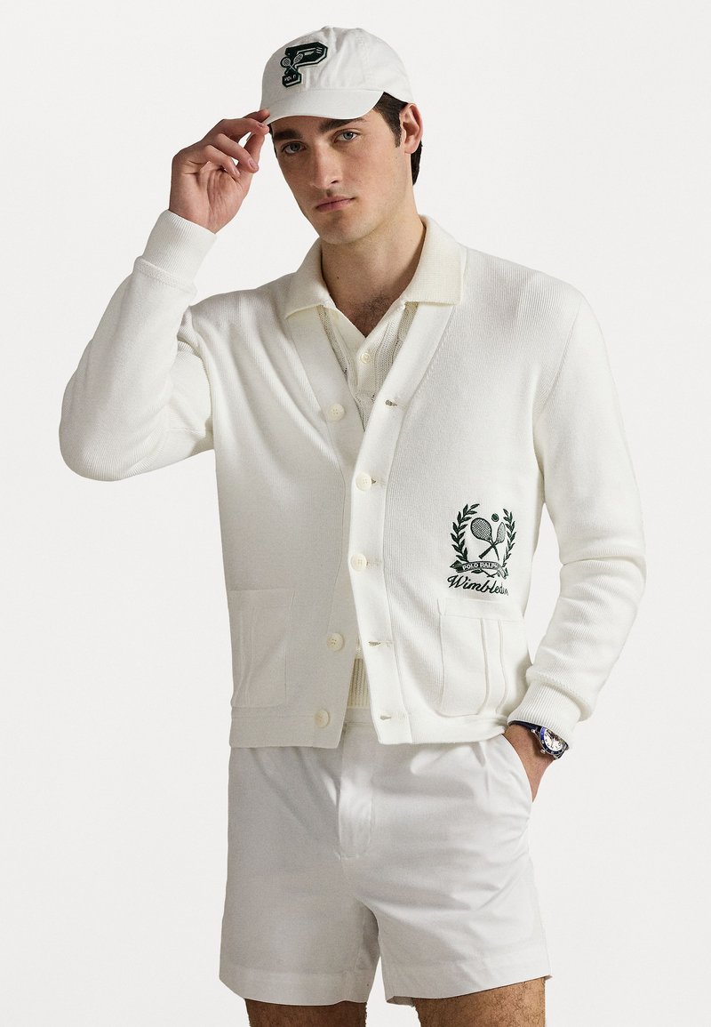 Cardigan blanc en mailles côtelées, avec un logo Wimbledon brodé en vert. La tenue comprend un polo blanc assorti et un short.