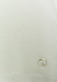 Lichtgroene katoenen stof met een gladde textuur; heeft een klein geborduurd logo in een iets donkerdere tint, geplaatst rechtsonder.