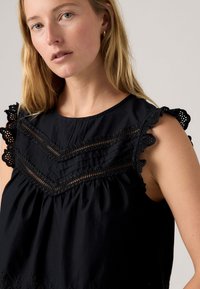 Blusa nera senza maniche con decorazione in pizzo sulle spalle e motivo a zigzag sul petto. Tessuto morbido e testurizzato con vita arricciata.