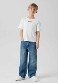 Witte katoenen t-shirt met zwarte tekst "Studio editie" op de borst, gecombineerd met lichtblauwe denim, wijde spijkerbroek en witte sneakers.