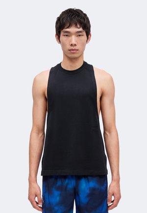 Ung mand iført en sort ærmeløs tanktop og blå shorts med mønster, står vendt mod kameraet foran en ensfarvet baggrund.