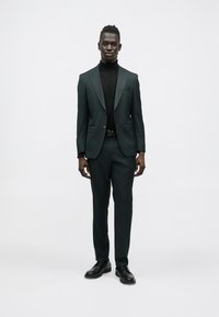 Fato verde escuro feito à medida com lapelas de entalhe, combinado com uma camisola de gola alta preta. Design ajustado e sapatos pretos polidos completam o visual.