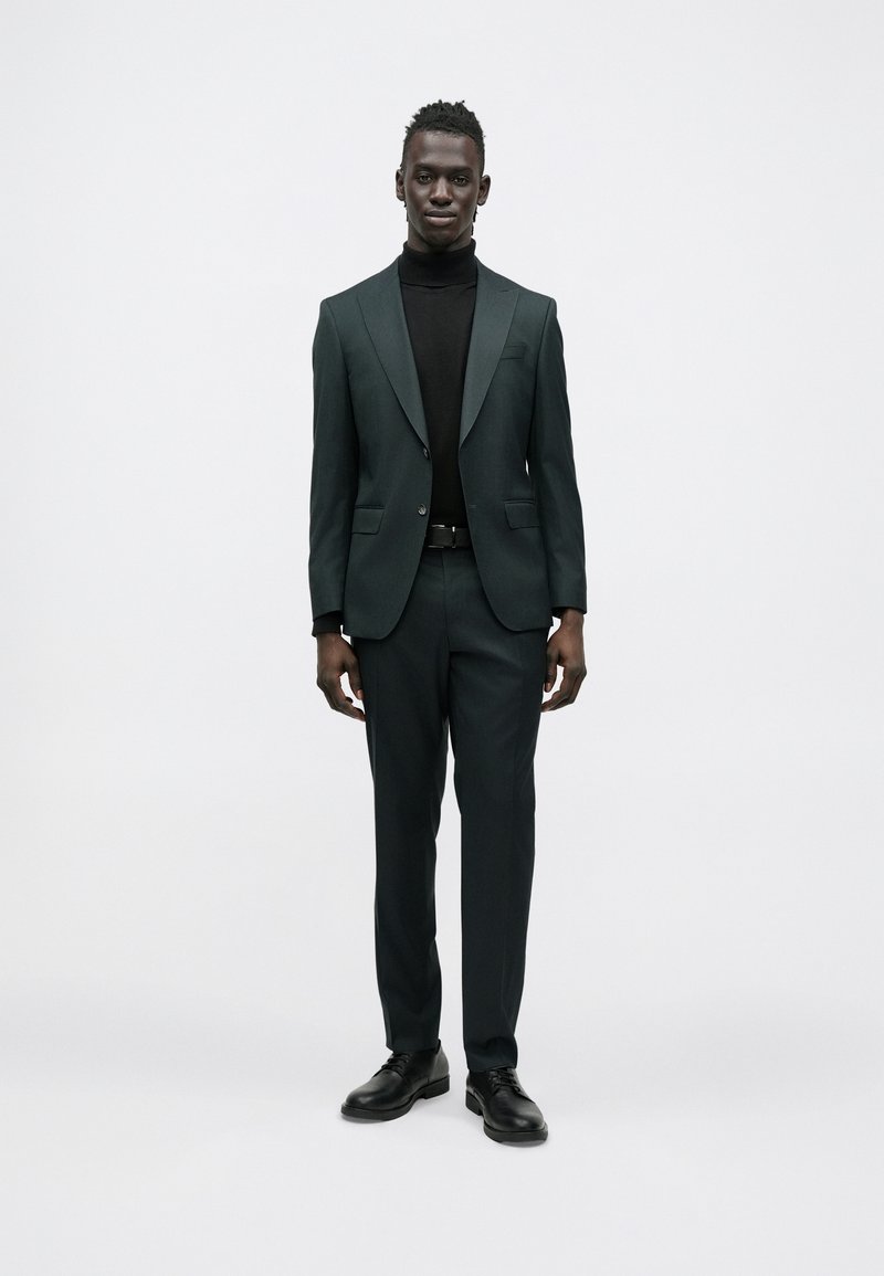 Fato verde escuro feito à medida com lapelas de entalhe, combinado com uma camisola de gola alta preta. Design ajustado e sapatos pretos polidos completam o visual.