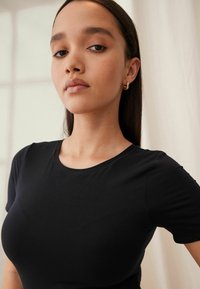 Zwarte fitted t-shirt met korte mouwen en een ronde halslijn, gemaakt van zachte stof. Het model heeft steil haar en draagt gouden creolen.