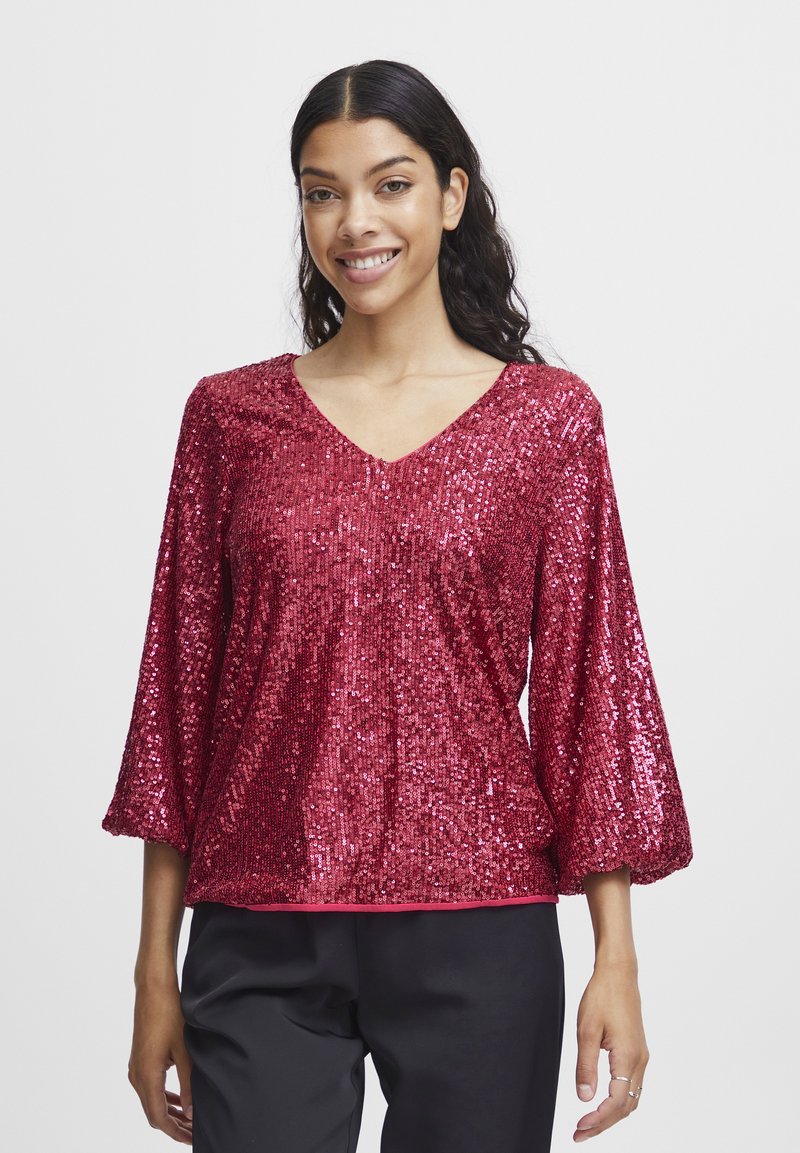 Top rosso con paillettes, scollo a V e maniche tre quarti. Texture morbida e vestibilità ampia, indossato con pantaloni neri.