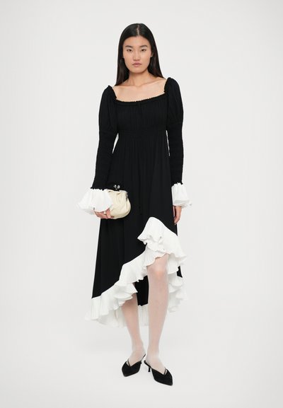 Rochie lungă neagră cu accente albe din volane, mâneci bufante, decolteu pătrat și corset ajustat; purtată cu o geantă mică de o culoare deschisă.