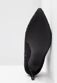 Escarpin noir à talon haut, avec une tige en tissu texturé ornée d'un design en dentelle complexe, un bout pointu et un talon stiletto mince et effilé.