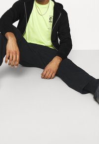 Felpa nera con zip, t-shirt grafica giallo neon, pantaloni neri e sneakers nere; posa seduta con le mani appoggiate sulle ginocchia.