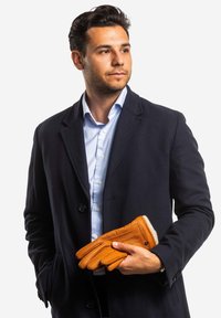 Leather gloves in orange with a ribbed knit cuff, held in a hand. Worn with a navy overcoat and light blue striped shirt.