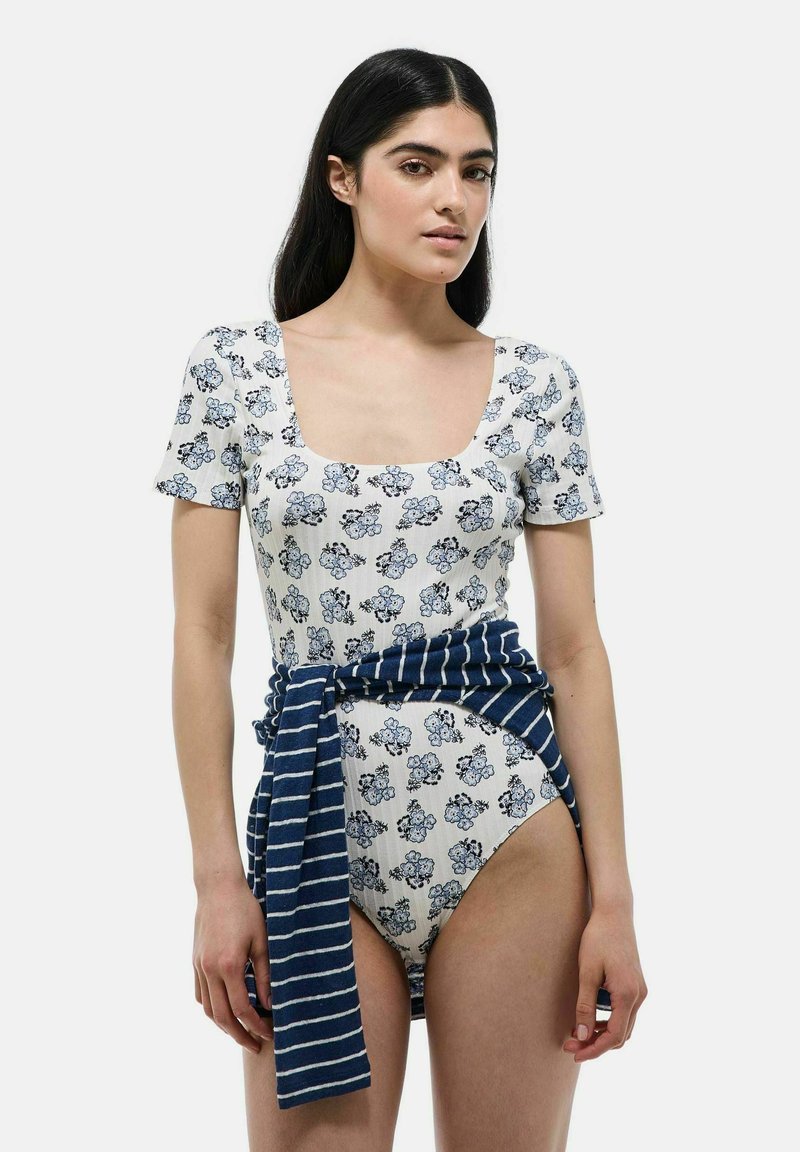 Maillot de bain blanc à motifs floraux avec décolleté carré et manches courtes, associé à un wrap rayé bleu marine à la taille. Texture lisse.