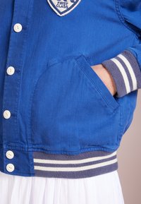 Modrá džínová bunda se zapínáním na bílý knoflík, kapsou na levé straně a pruhovanými manžetami v námořnické modré a bílé. Obsahuje nášivku "FIRST CLASS".