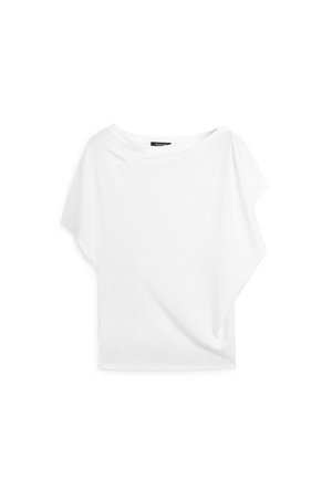 Blouse blanche asymétrique à manches courtes avec un large col et un détail en tissu drapé d’un côté, présentée sur un fond uni.