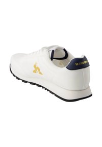 Sneaker bianca con tomaia in pelle liscia, accento del logo dorato e linguetta del tallone in blu navy. Suola in gomma nera con un motivo di battistrada testurizzato.