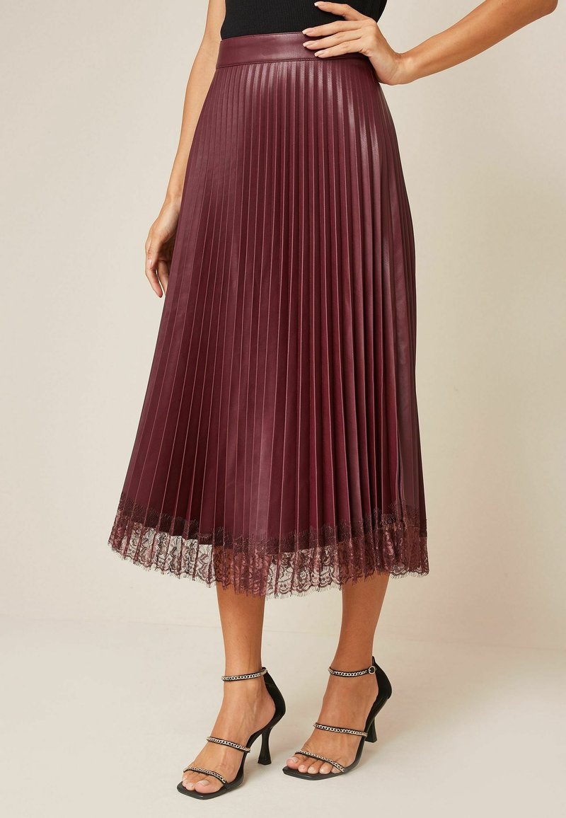 Femme portant une jupe midi plissée bordeaux avec un ourlet en dentelle, associée à des sandales noires à talons hauts et fines brides, debout devant un fond neutre.