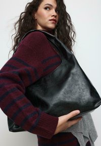 Črna usnjena torbica za čez ramo z gladko teksturo, širokim odprtjem in zaobljeno obliko, držana ob bordo in sivo pleteno pulover.