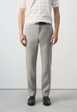 Hombre que lleva pantalones de vestir gris claro, cinturón negro, mocasines negros y una camisa blanca de manga corta con finas rayas horizontales.