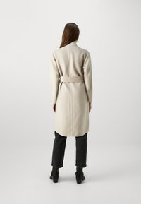 Beige villasegu segu coat, millel on vöödega talje, pikad varrukad ja sirge lõige, koos tumedate teksade ja kingadega.