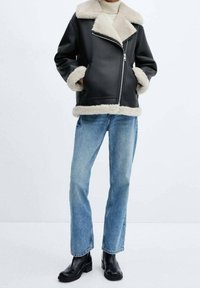 Veste en cuir noire avec col et poignets en shearling beige, fermeture éclair, associée à un jean droit bleu clair et des bottines noires.