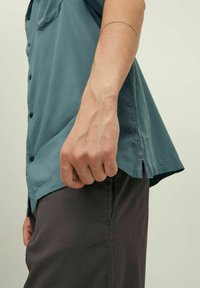 Braccio maschile con un tatuaggio minimale vicino al gomito e un tatuaggio su un dito, che indossa una maglietta a maniche corte color turchese e pantaloni grigio scuro, su uno sfondo neutro.