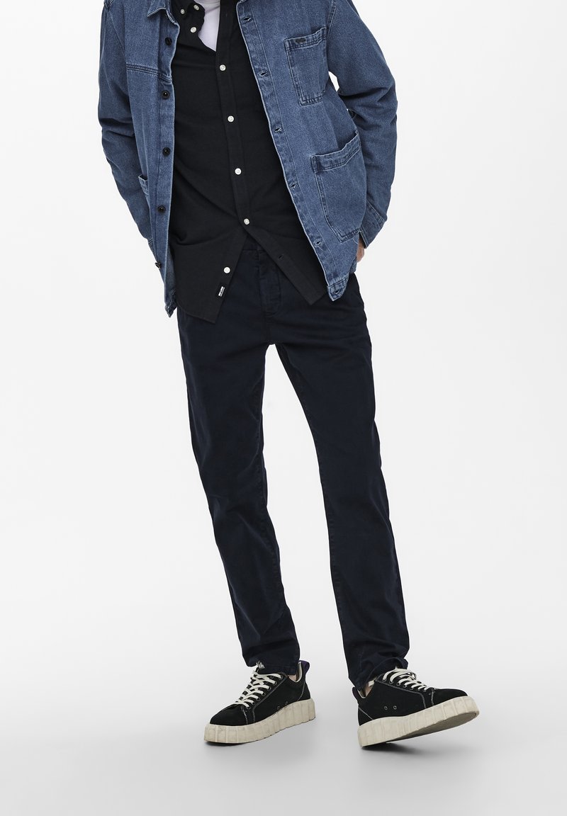 Giubbotto di jeans blu, camicia nera sotto, pantaloni scuri e sneakers nere con suola beige. Outfit casual a strati.