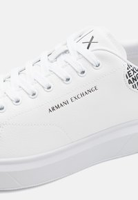 Zapatilla blanca hecha de cuero sintético texturizado. Presenta cordones blancos, una suela blanca elevada y el logo "Armani Exchange" en negro.