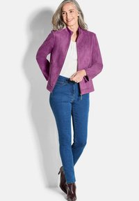 Veste en suede violette avec un col montant et des poches zippées, portée sur un haut blanc et un jean bleu. Le mannequin pose avec un léger sourire.