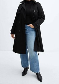 Trench-coat noir avec une fermeture à un seul bouton, associé à un jean bleu clair à jambes larges et des bottes noires à bout pointu. Tissu lisse et design classique.